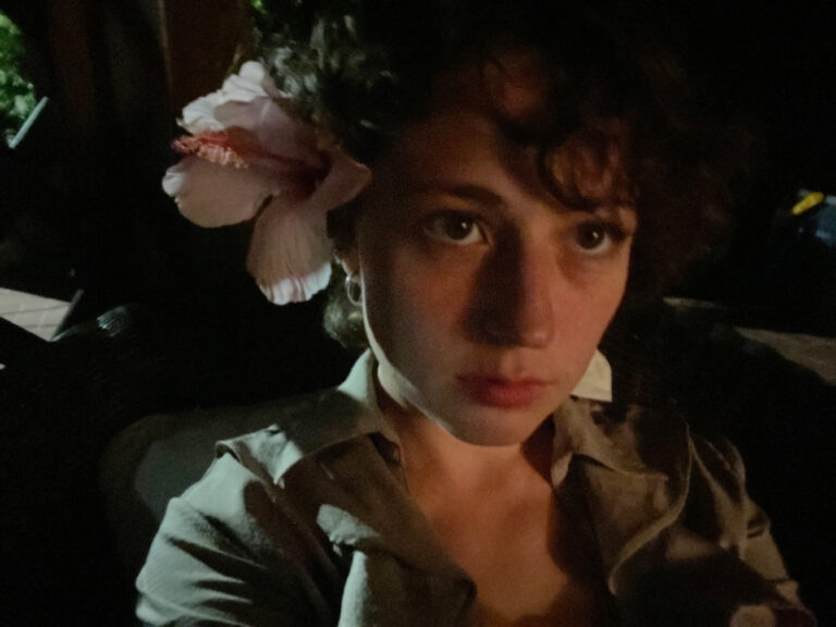 A portrait of a young woman with curly hair and a large pink flower in her hair, capturing a moody and stylish aesthetic with low lighting, perfect for fashion or lifestyle SEO.